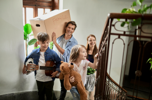 A nuclear family climbing up to the second floor of their new home. The dad is carrying a box on his shoulder that says "fragile."